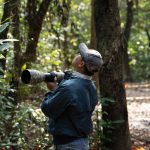 Duas pessoas observam aves em uma trilha de floresta usando câmeras com lentes teleobjetivas.