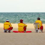 Três guarda-vidas uniformizados observam o mar sentados na areia da praia.