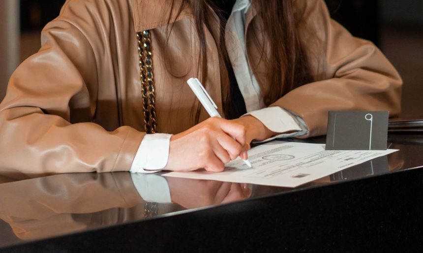 Mulher assina um documento em um balcão de atendimento administrativo.