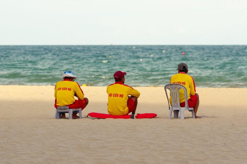 Três guarda-vidas uniformizados observam o mar sentados na areia da praia.