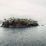 Vista costeira da Ilha das Cabras, em Ilhabela, com embarcações no mar e vegetação ao redor.